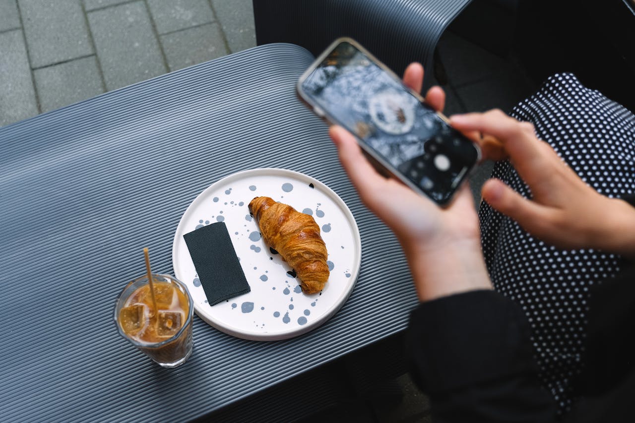 trusted-partner Close-up of a croissant and iced coffee on a terrace table, captured with a smartphone.